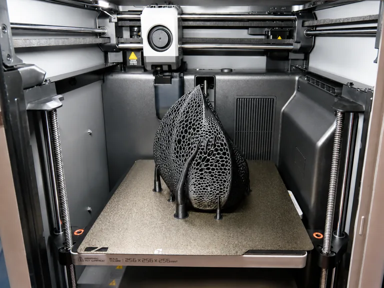 A black 3D-printed shade component being printed inside a desktop 3D printer