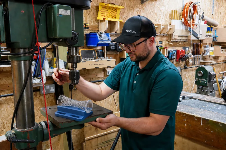 Sami drilling a crystal vase on a pillar drill using a custom blue V-block jig to safely support the vase during the drilling process.